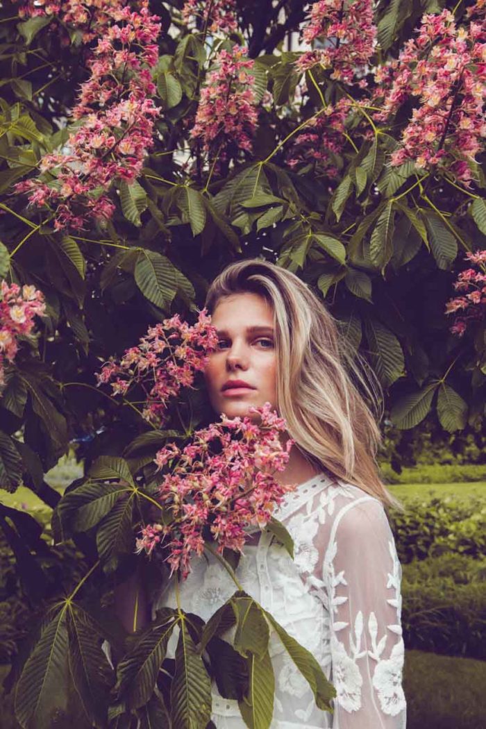 Blonde model in white dress and pink lipstick behind pink lilac tree